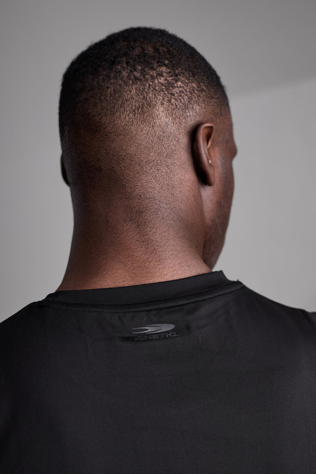 Back view of a person wearing a black shirt with a visible brand logo on a plain background