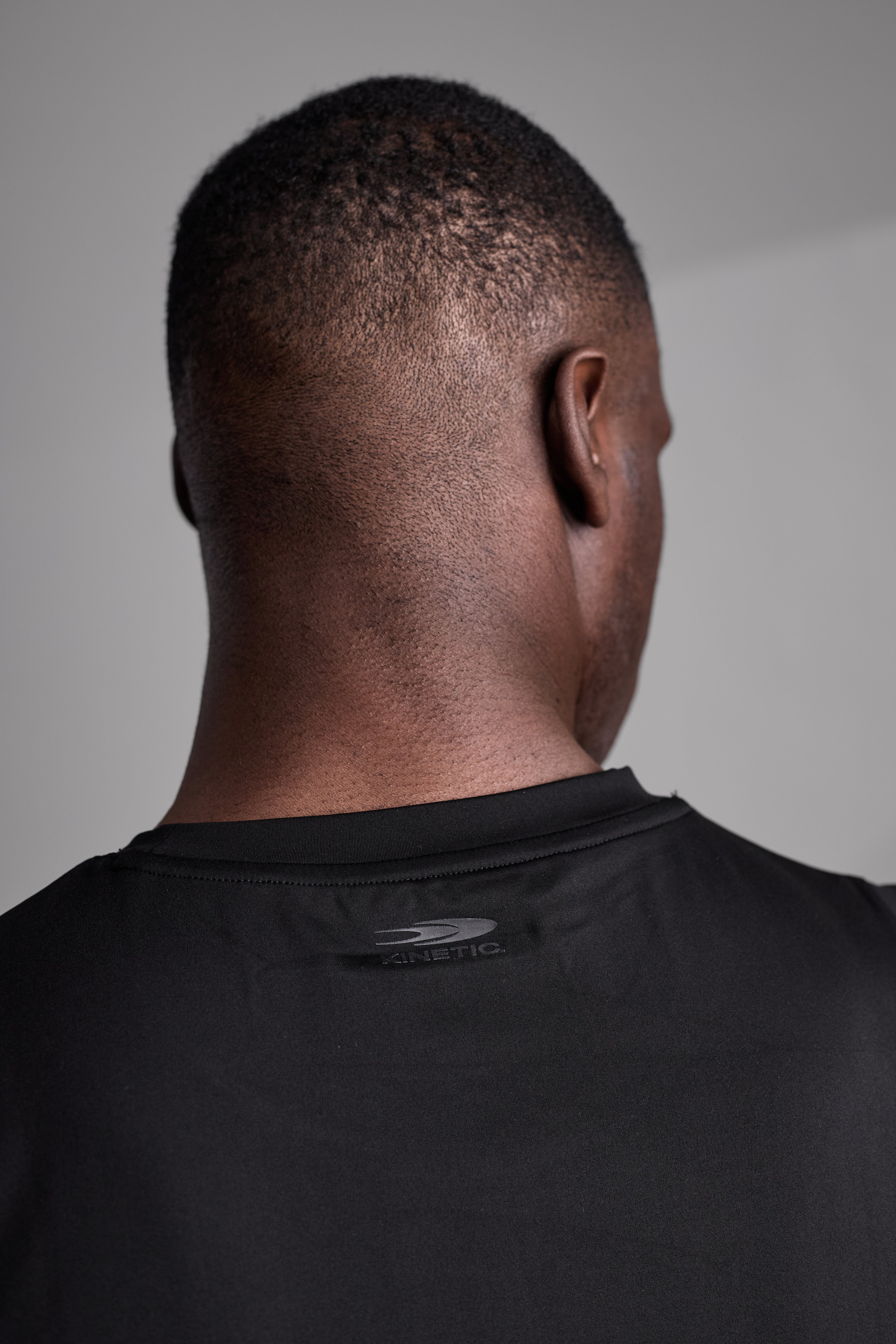 Back view of a person wearing a black shirt with a visible brand logo on a plain background