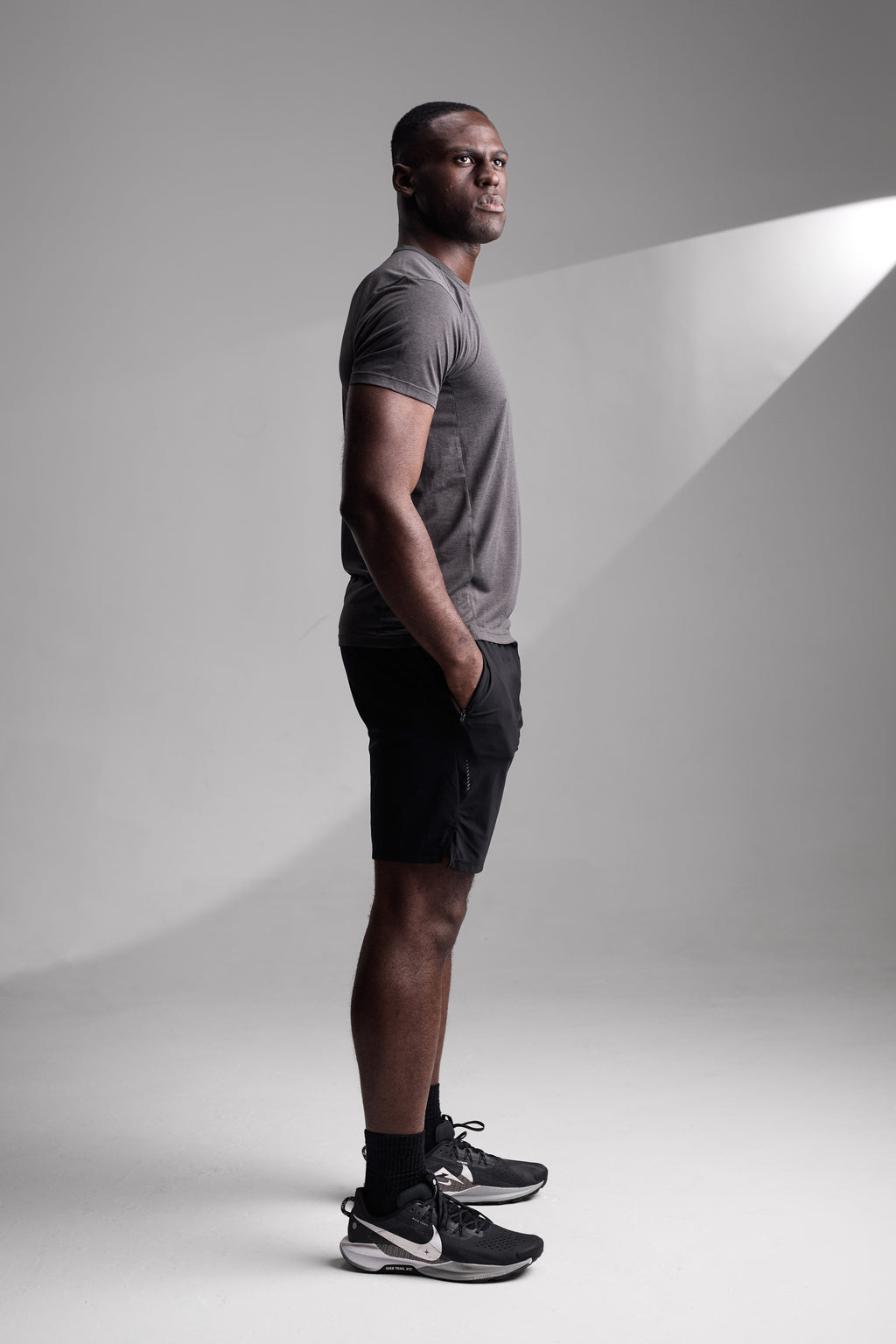Man standing side-on wearing a gray fitness t-shirt, black shorts, and black sneakers on a plain background